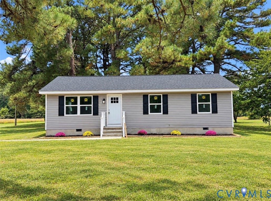 Single-story residence featuring grey vinyl siding, black louvered shutters, shingled roof, front entry with white door, and mature evergreen trees Single-story residence featuring grey vinyl siding, black louvered shutters, shingled roof, front entry with white door, and mature evergreen trees
