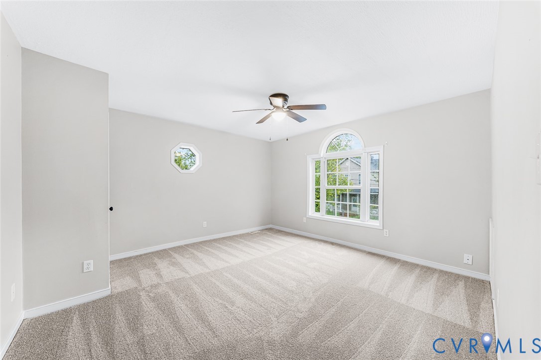 Neutral-toned interior featuring wall-to-wall carpeting, an arched window, a secondary octagonal window, and a central ceiling fan