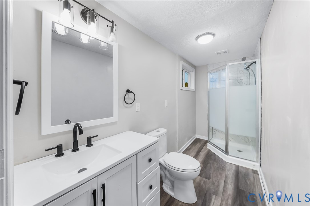 White vanity with a rectangular sink, matte black hardware, wood-finish flooring, and a glass-enclosed shower with a rainfall showerhead