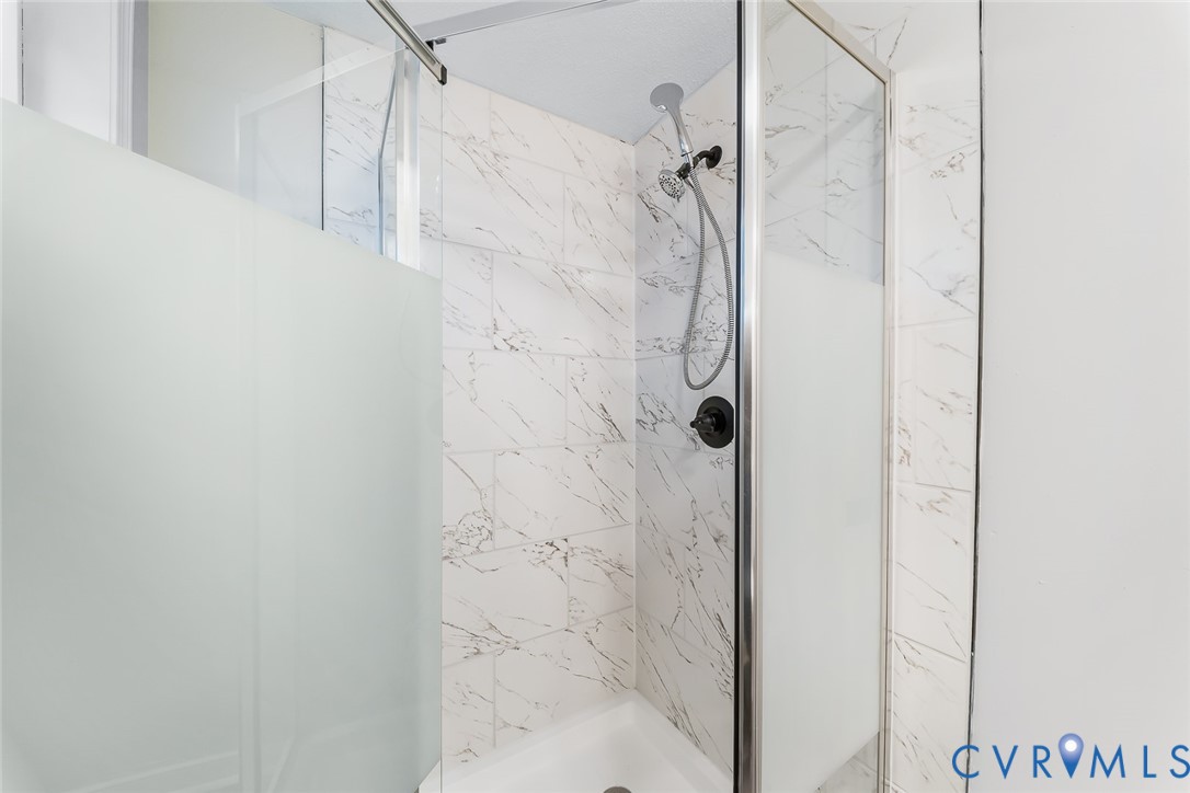 Walk-in shower enclosure featuring large-format patterned wall tile, sliding frosted glass doors, chrome frame, and a wall-mounted handheld shower fixture with dark finish hardware