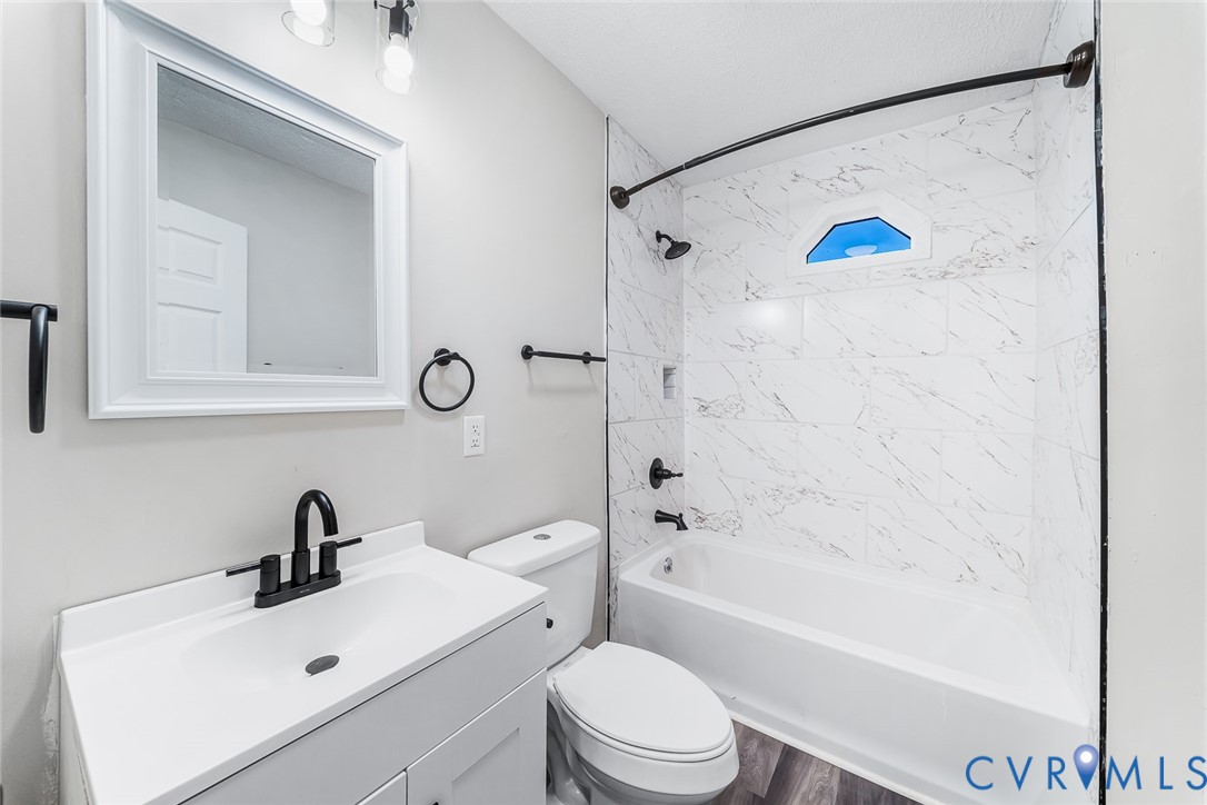 Bathroom featuring a white vanity cabinet, rectangular undermount sink with matte black faucet, wall-mounted rectangular mirror, and tub-shower surround with light-toned stone-finish tile and matte black hardware