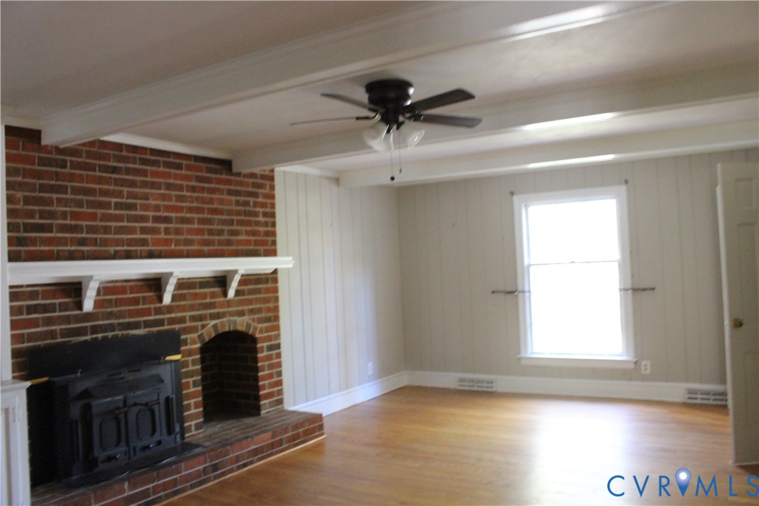 Exposed ceiling beams, brick fireplace featuring a wood-burning insert and mantel, wood-finish flooring, and a ceiling fan with integrated lighting