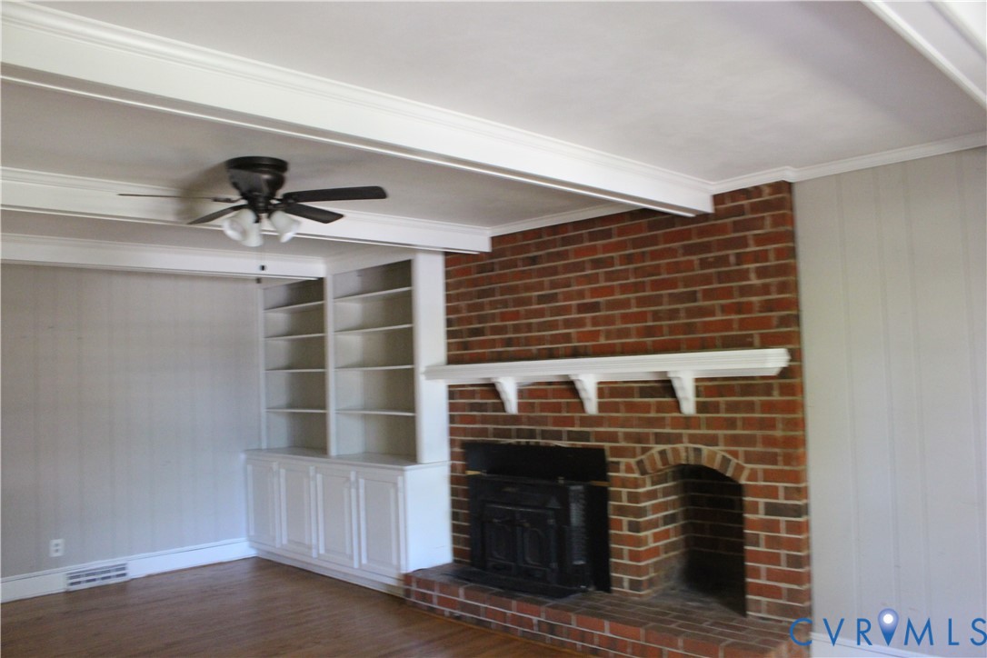 Floor-to-ceiling red brick fireplace with an attached mantel, wood-finish flooring, exposed ceiling beams, and built-in white cabinetry with integrated shelving