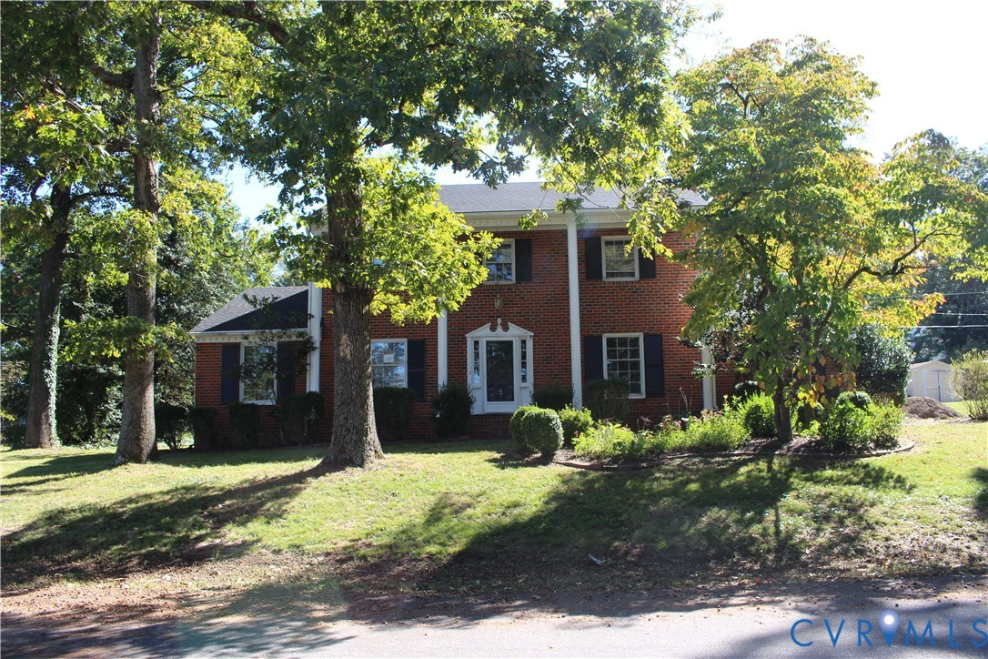 Two-story brick exterior featuring classic white columns, a covered entryway, decorative dark shutters, and a mature tree-lined landscape