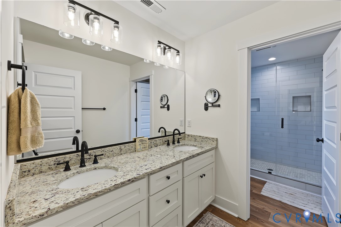 Double vanity with stone countertop, undermount sinks, matte black fixtures, large frameless mirror, and shower enclosure with subway tile surround