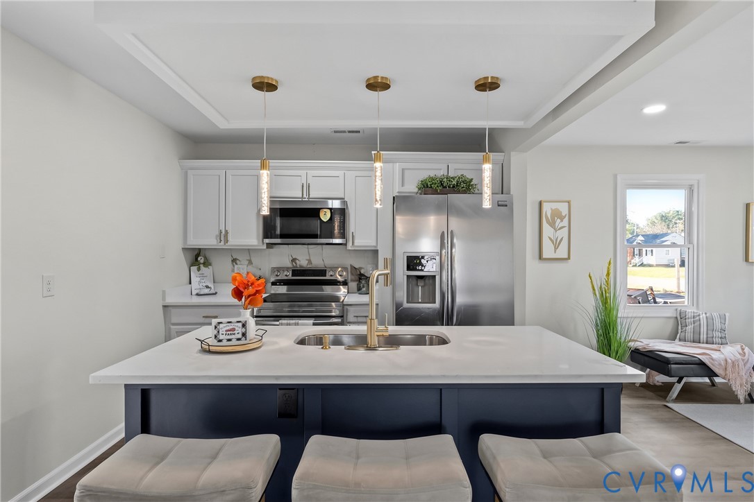 Kitchen featuring a central island with a solid-surface countertop and undermount sink, white cabinetry, stainless steel appliances, recessed ceiling detail, and three glass pendant lights Kitchen featuring a central island with a solid-surface countertop and undermount sink, white cabinetry, stainless steel appliances, recessed ceiling detail, and three glass pendant lights