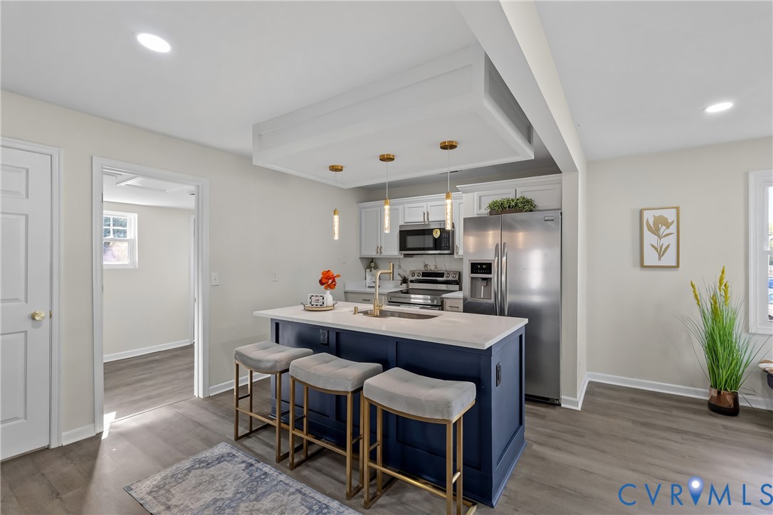 Kitchen featuring a central island with a solid surface countertop, contrasting navy blue cabinetry, stainless steel appliances, recessed ceiling lighting, and wood-finish flooring Kitchen featuring a central island with a solid surface countertop, contrasting navy blue cabinetry, stainless steel appliances, recessed ceiling lighting, and wood-finish flooring