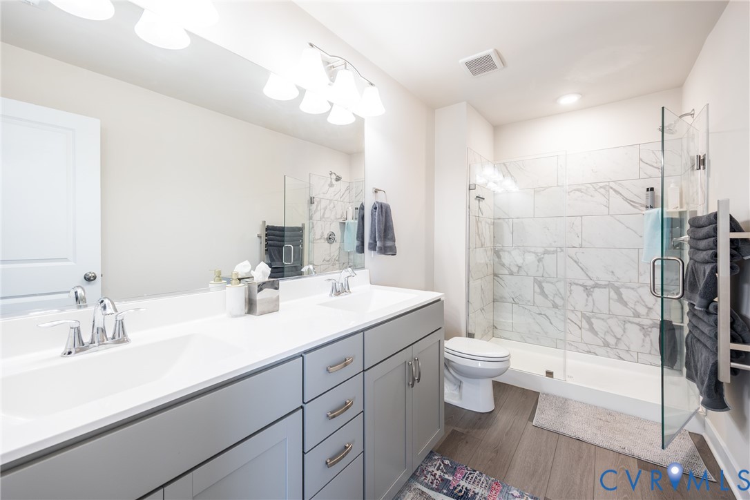 Dual-sink vanity with white stone countertop, grey shaker-style cabinetry, walk-in shower with floor-to-ceiling tile surround, glass enclosure, and wood-finish flooring Dual-sink vanity with white stone countertop, grey shaker-style cabinetry, walk-in shower with floor-to-ceiling tile surround, glass enclosure, and wood-finish flooring