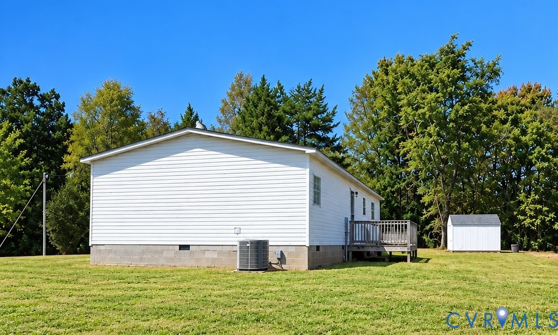 White vinyl siding exterior featuring a block foundation, outdoor wood deck, central air conditioning unit, and a detached storage shed situated on a grass lot White vinyl siding exterior featuring a block foundation, outdoor wood deck, central air conditioning unit, and a detached storage shed situated on a grass lot