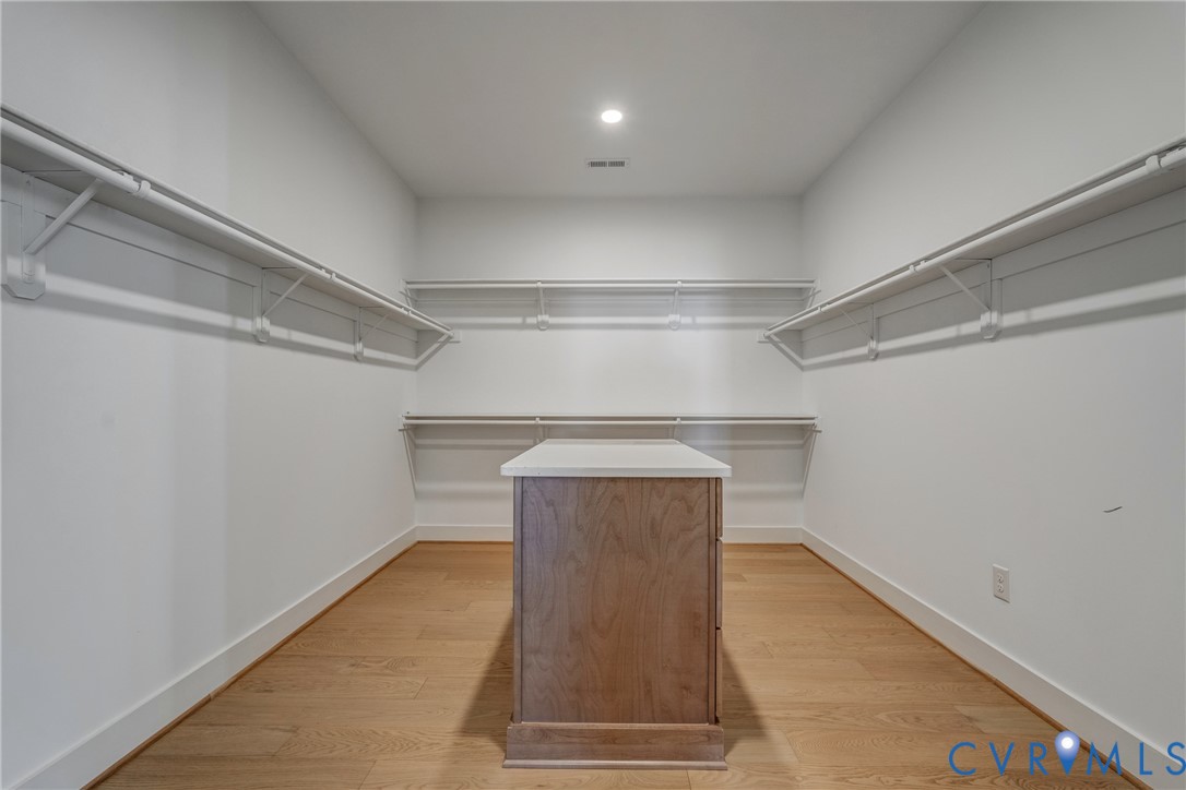 Walk-in storage space featuring wood-finish flooring, white shelving tiers, lighting, and a central island with a solid surface countertop