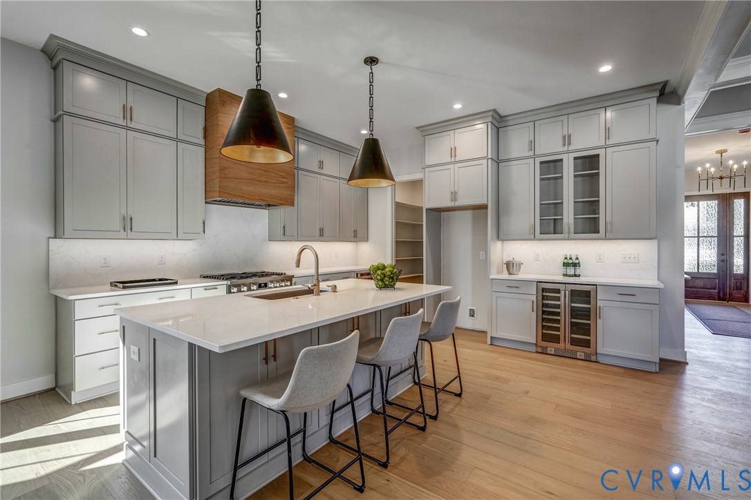 Kitchen featuring shaker-style cabinetry, an oversized quartz-topped island, wood-finish flooring, a custom wood-paneled range hood, and integrated stainless steel beverage refrigeration Kitchen featuring shaker-style cabinetry, an oversized quartz-topped island, wood-finish flooring, a custom wood-paneled range hood, and integrated stainless steel beverage refrigeration