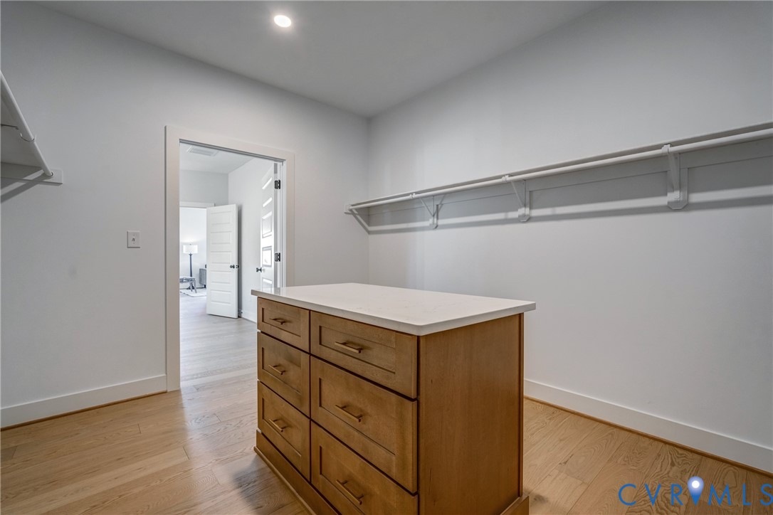 Walk-in closet featuring wood-finish flooring, a central island with a stone-top surface and six drawers, and wall-mounted shelving with hanging rods