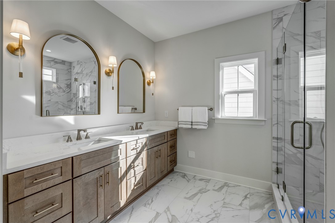 Double vanity with wooden cabinetry and stone countertop, dual arched wall mirrors, wall-mounted brass light fixtures, glass-enclosed shower with tiled surround, and large-format tile flooring Double vanity with wooden cabinetry and stone countertop, dual arched wall mirrors, wall-mounted brass light fixtures, glass-enclosed shower with tiled surround, and large-format tile flooring