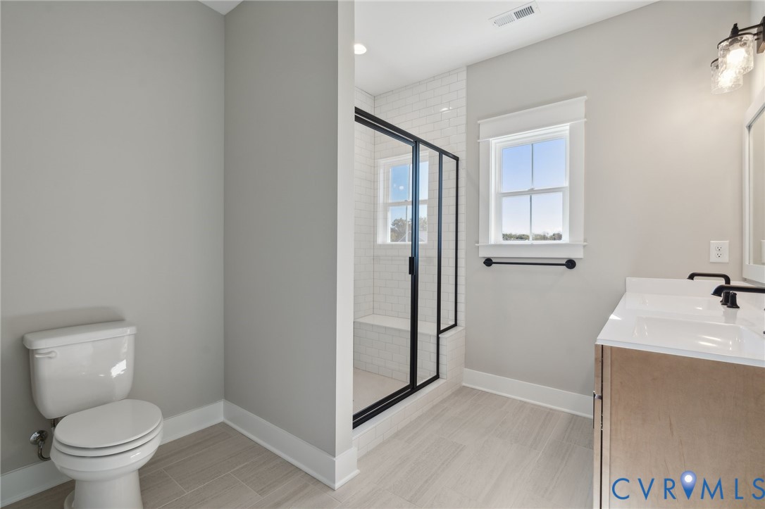 Walk-in shower with white subway tile surround, glass enclosure with black metal frame, double vanity with solid surface countertop, and wood-finish flooring