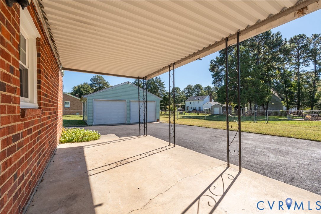Covered concrete patio featuring decorative metal support posts, brick exterior, paved driveway, and detached two-car garage