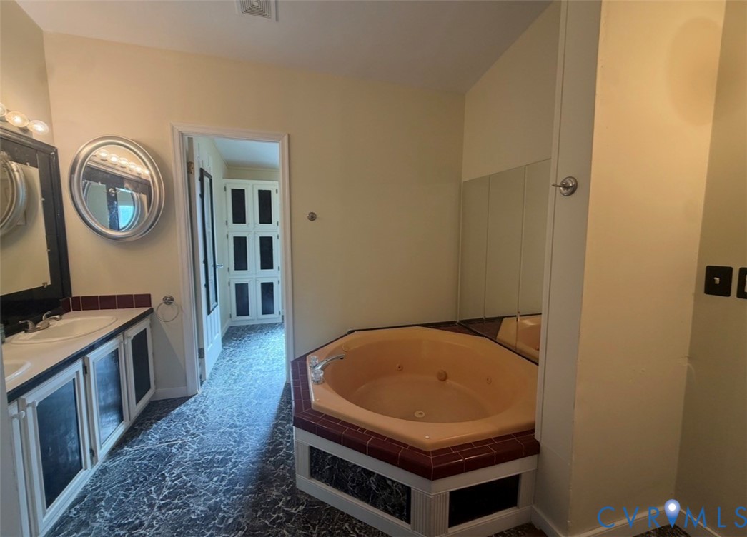 Bathroom featuring a dual-sink vanity with dark-toned cabinetry, circular wall mirrors, a drop-in jetted tub with tiled surround, and floor-to-ceiling cabinetry