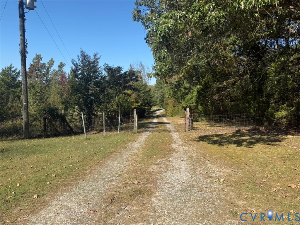 Gravel driveway flanked by perimeter wire fencing, wooden gate posts, and mature tree lines