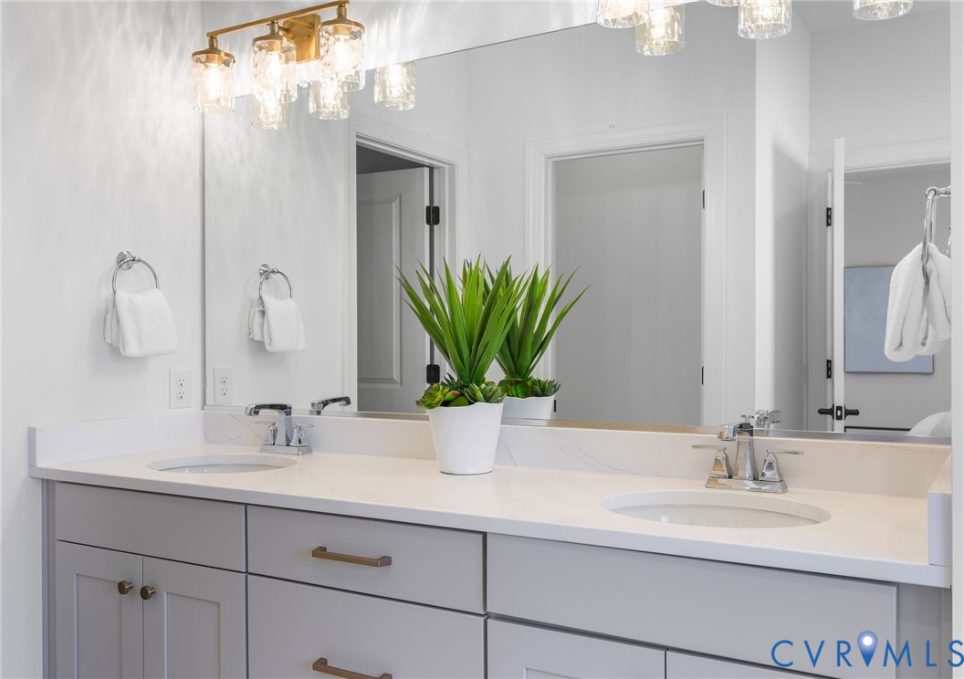 Dual-sink vanity with a solid-surface countertop, light gray shaker-style cabinetry, metallic hardware, and a wall-to-wall mirror illuminated by multi-light glass fixtures