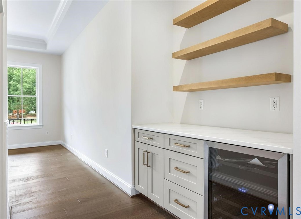 Built-in cabinetry featuring light-toned shaker-style doors, matte hardware, quartz countertop, integrated wine refrigerator, and floating wood-finish shelving, complemented by wood-finish flooring and crown molding