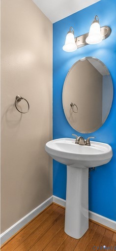 Pedestal sink with polished metal dual-handle faucet, oval frameless wall mirror, brushed nickel dual-light fixture, and circular towel ring, accented by wood-finish flooring