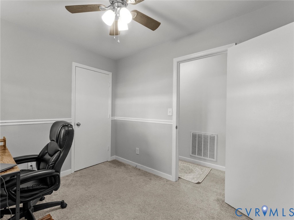 Neutral-toned interior featuring wall-mounted chair rail molding, wall-to-wall carpeting, and a ceiling-mounted light fixture with a multi-blade fan