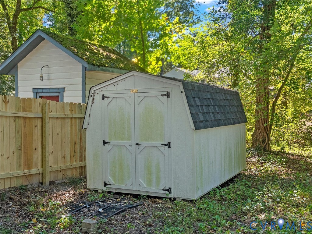 Gable-roof storage structure featuring double-door entry and asphalt shingle roofing, situated within a wooded lot with wood-panel privacy fencing