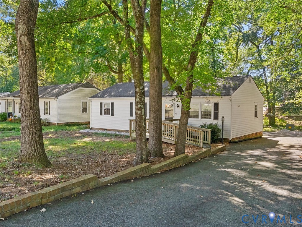 Exterior featuring white siding, a paved driveway, a stepped masonry retaining wall, a wood deck with railings, and mature trees