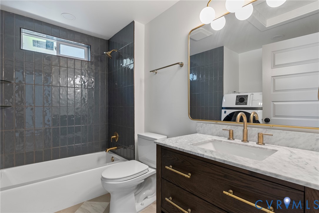 Dark wood vanity with a stone countertop, brass hardware, oversized gold-framed mirror, and vertical charcoal tile bathtub surround