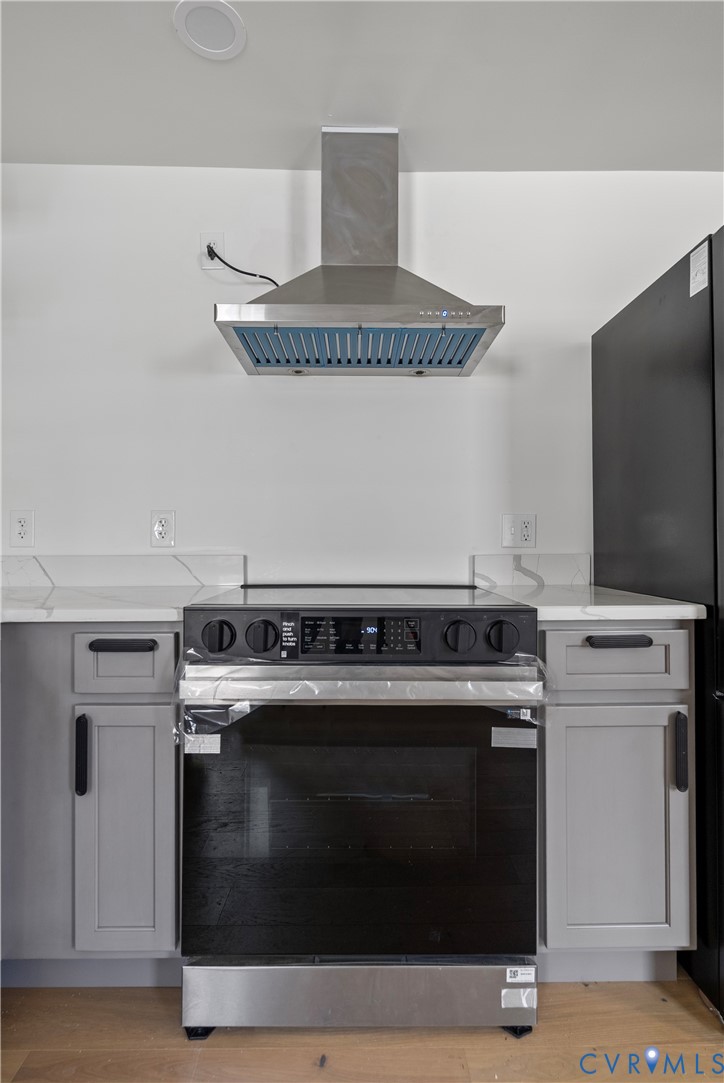 Stainless steel chimney-style range hood, electric slide-in range, stone-composite countertop with matching backsplash, and grey shaker-style cabinetry with matte black hardware