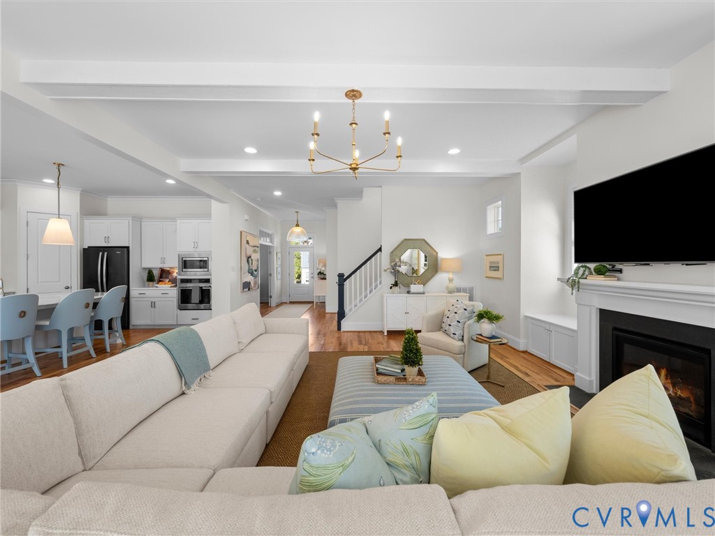 Open-concept interior featuring beamed ceilings, white shaker-style cabinetry, wood flooring, a fireplace with a mantel, and a multi-arm chandelier