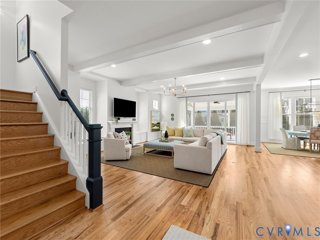 Open-concept floor plan featuring wood-finish flooring, recessed lighting, exposed ceiling beams, a built-in fireplace, and a staircase with a contrasting dark-finish handrail