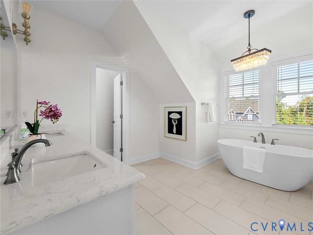 Freestanding soaking tub with a deck-mounted faucet, rectangular undermount sink, stone-surface countertop, vaulted ceiling, and tiered crystal chandelier