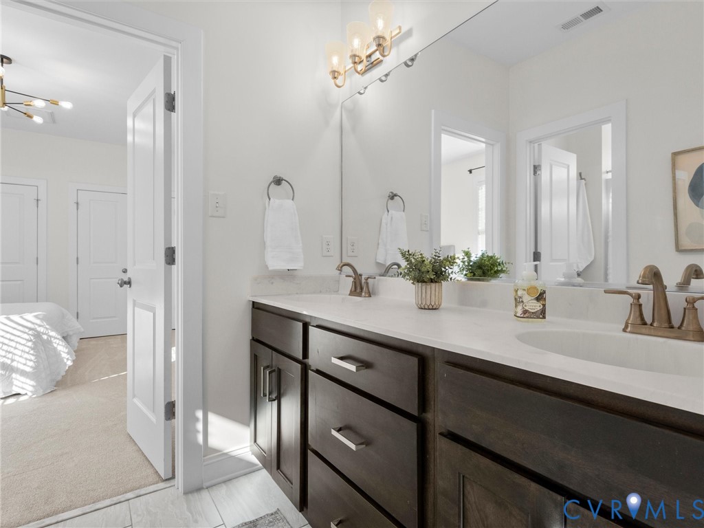 Dark wood cabinetry featuring shaker-style doors, stone-surface countertop with dual undermount sinks, bronze-tone finish faucets, and a large-format vanity mirror with integrated three-bulb light fixture