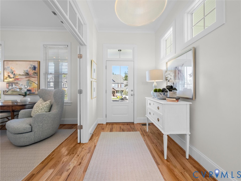 Foyer featuring wood-finish flooring, a glass-panel entry door with transom window, double-hung windows, crown molding, and a drum-style ceiling fixture Foyer featuring wood-finish flooring, a glass-panel entry door with transom window, double-hung windows, crown molding, and a drum-style ceiling fixture