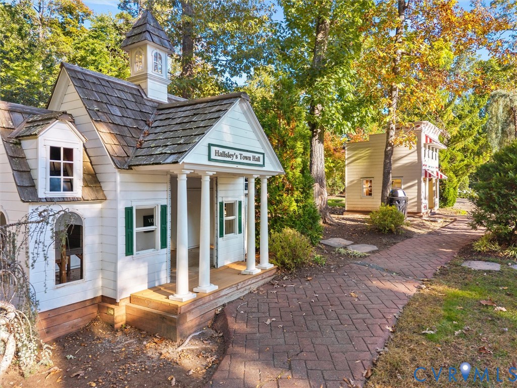 Miniature exterior structure featuring white horizontal siding, a cedar-shake roof with a cupola, a front portico with classical columns, and herringbone-patterned brick hardscaping