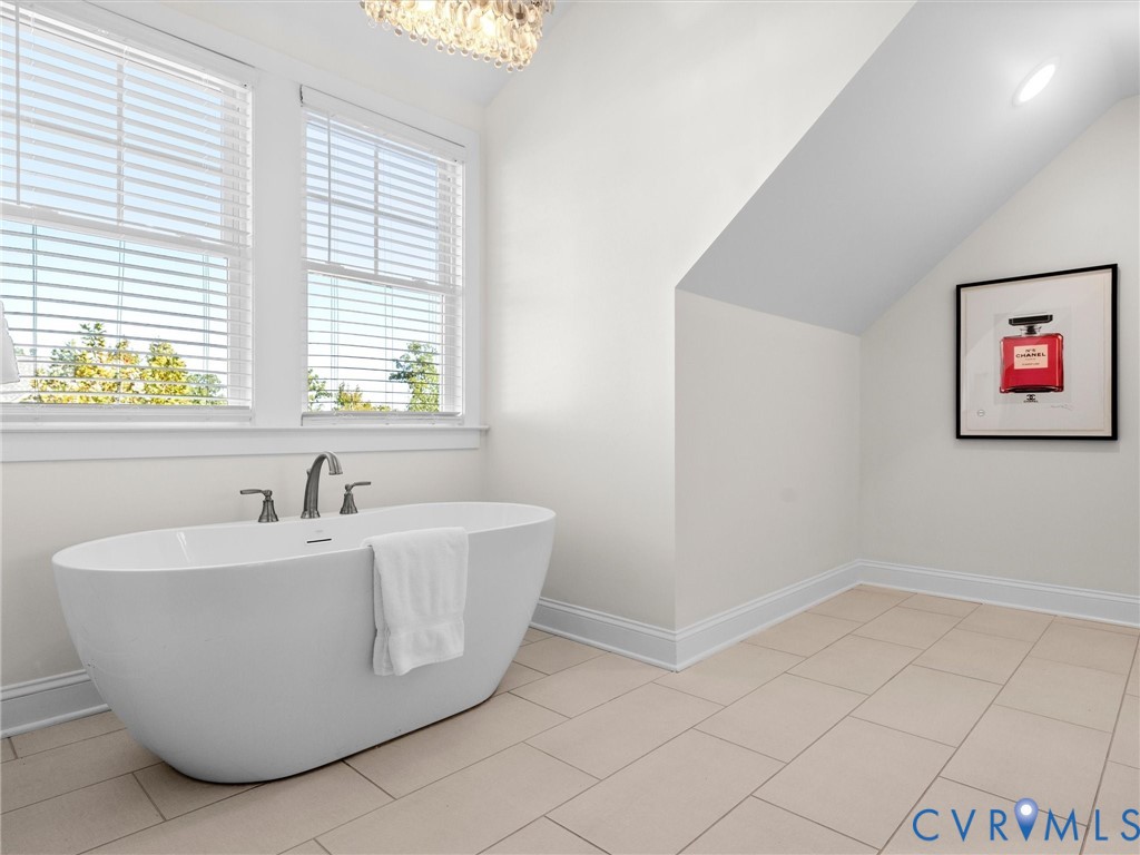 Freestanding white soaking tub with deck-mounted chrome faucet, large-format tile flooring, double windows, and recessed ceiling lighting