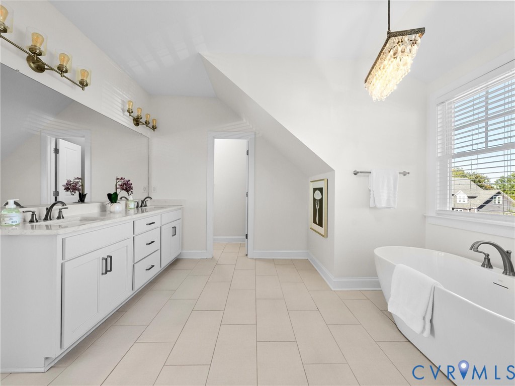 Dual-sink vanity with stone countertop, freestanding soaking tub, large-format tile flooring, rectangular wall mirror, and crystal chandelier fixture