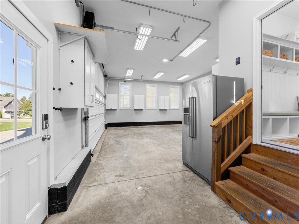 Garage space featuring white wall-mounted storage cabinetry, overhead fluorescent lighting, a stainless steel refrigerator, and wooden staircase leading to the interior