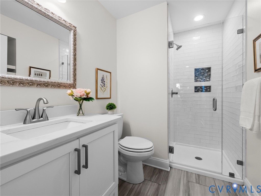 White vanity with stone countertop and metal hardware, subway tile shower enclosure with integrated shelving, glass shower door, and wood-finish flooring