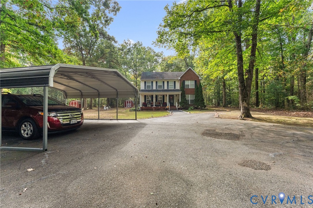 Two-story brick exterior home featuring a covered front porch, an attached metal carport, and a gravel driveway situated on a wooded lot