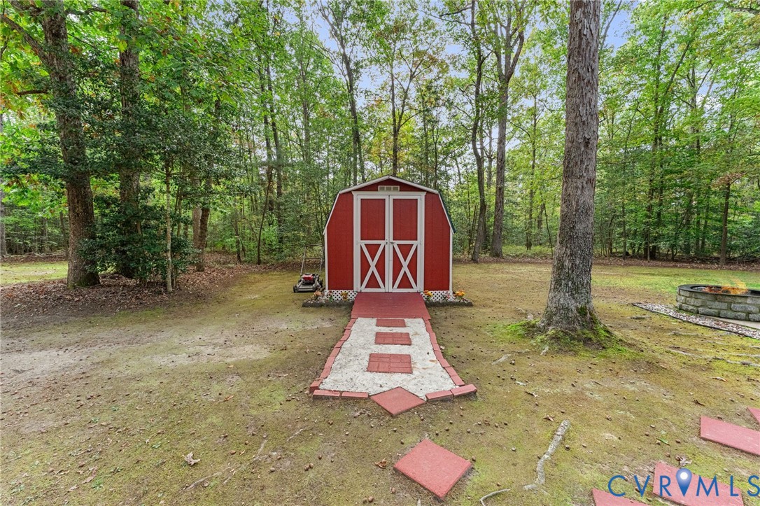 Storage structure featuring a gambrel roof, red-painted exterior with white trim, double-door entry, gravel and paver pathway, and a stone-built fire pit
