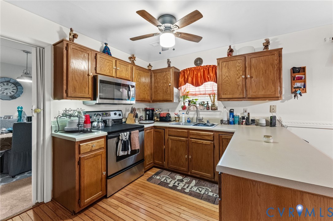 Kitchen featuring wood cabinetry, stainless steel appliances, neutral countertops, wood-finish flooring, and a ceiling fan with integrated lighting