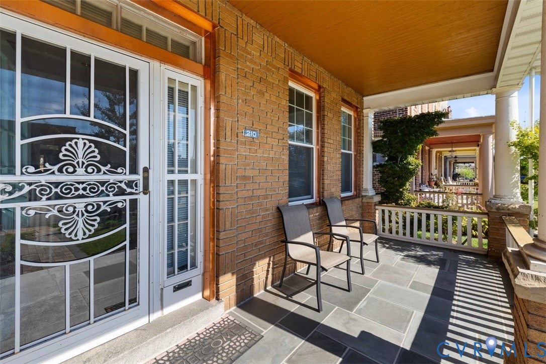 Covered front porch featuring a stone tile floor, brick exterior wall, decorative metal security door, and classic columns