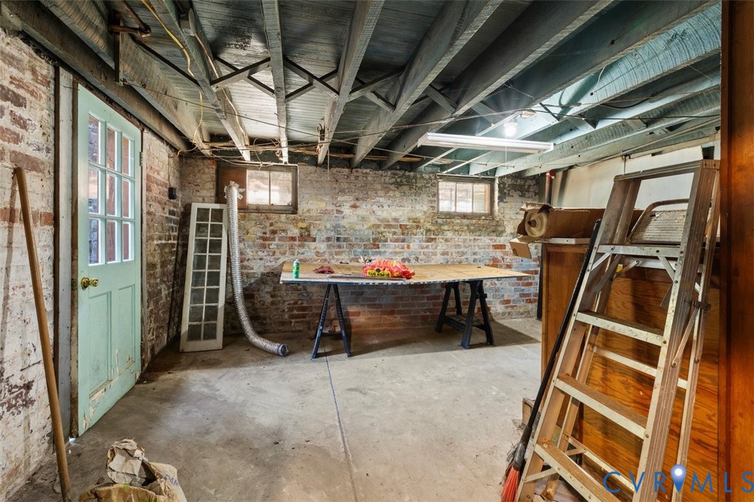 Lower-level space featuring exposed wood joist ceilings, painted brick masonry walls, concrete flooring, and an exterior-access door with glass lites