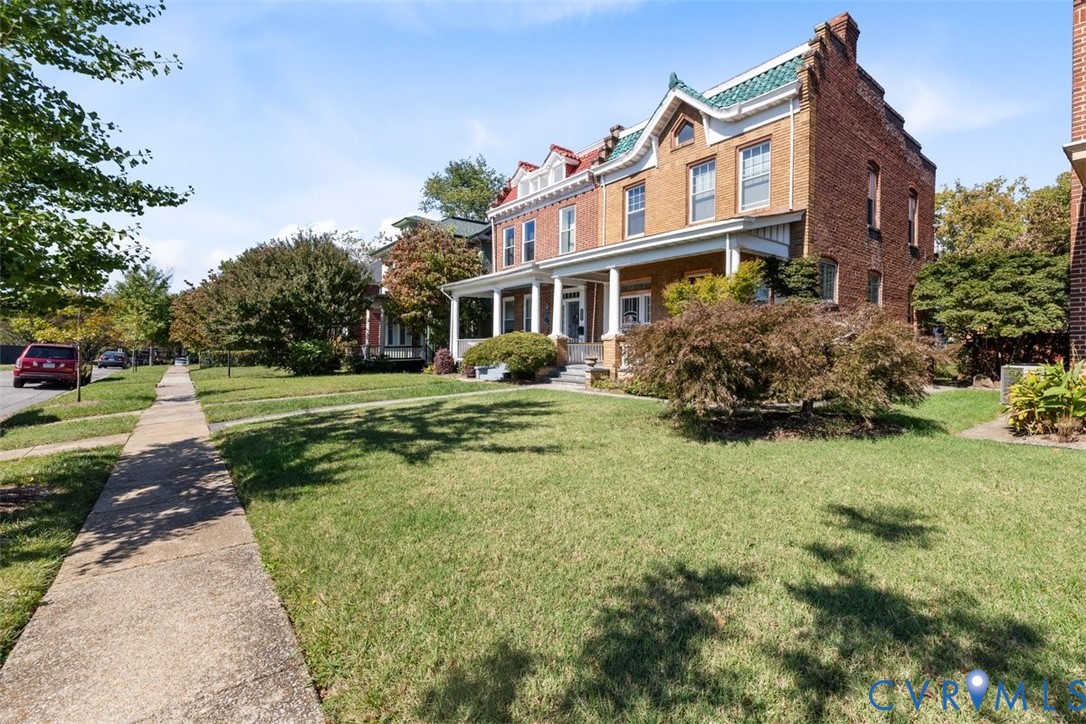 Traditional brick exterior featuring a covered front porch, dual dormer windows, green shingle roof accents, and mature landscaping