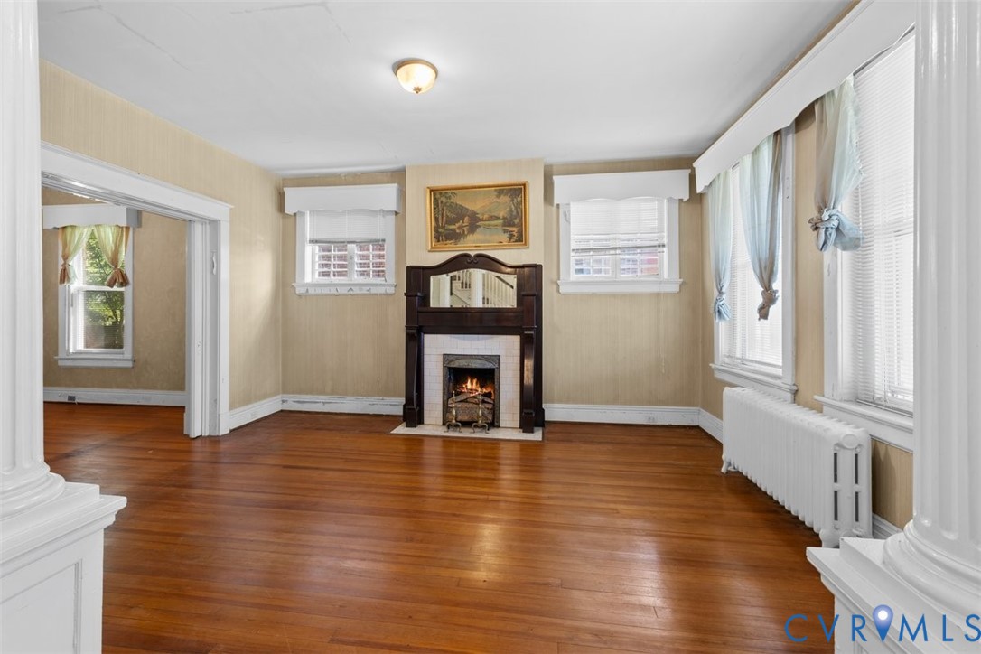 Living area featuring original wood flooring, a central fireplace with an ornate dark wood mantel, decorative column accents, and multiple windows with white trim