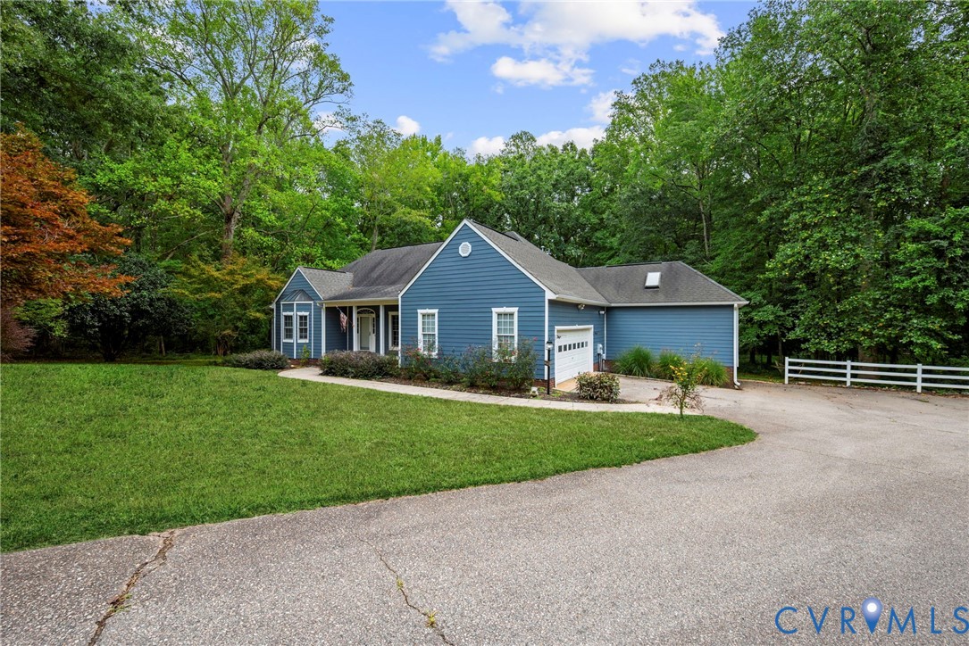 Blue-sided exterior featuring an asphalt shingle roof, attached garage, manicured lawn, and white rail fencing at the end of the driveway. Virtually edited grass