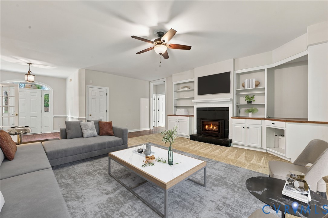 Living area featuring a fireplace with black brick, built-in cabinetry with shelving, a ceiling fan, and a front entry door with a glass transom. Virtually staged concept