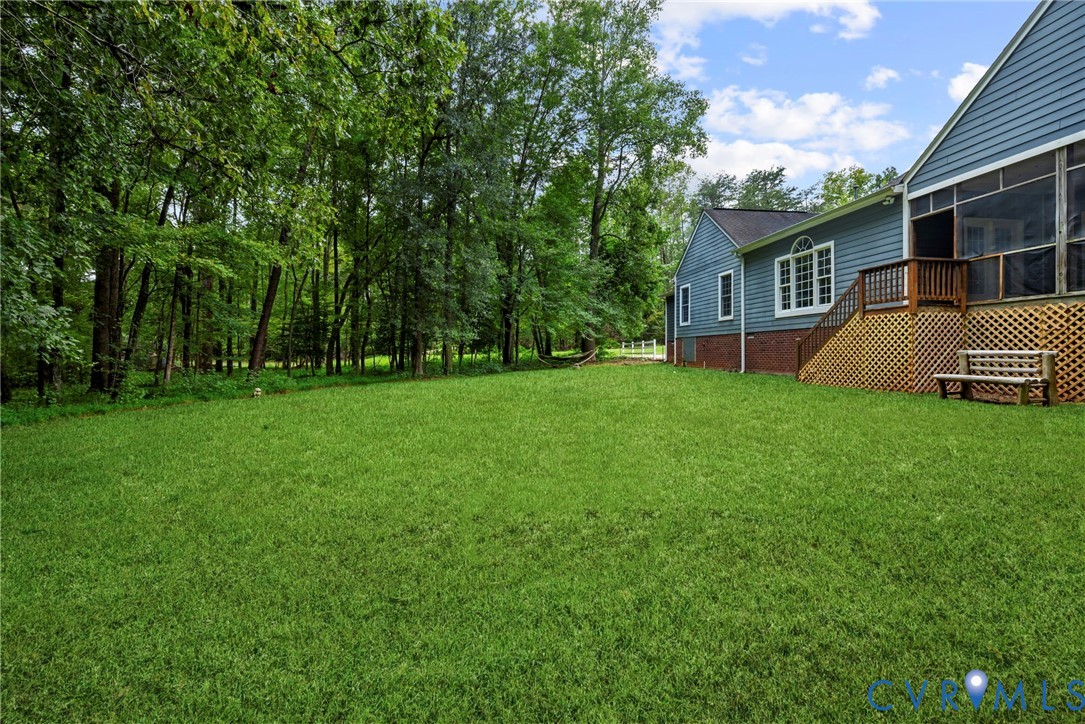 Expansive backyard featuring a level lawn, mature trees, blue-toned siding, brick foundation, and an elevated screened porch. Virtually edited grass