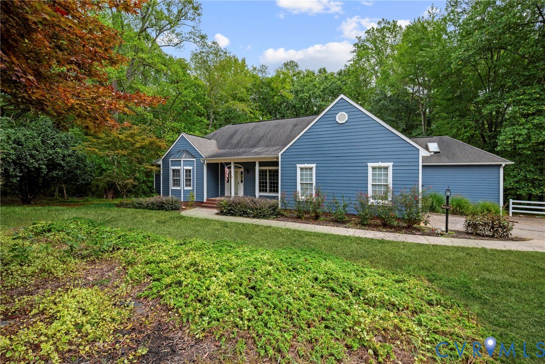 Blue siding exterior featuring a gable roofline, multi-pane windows, a recessed front entrance with a covered porch, and landscaped grounds with mature trees. Virtually edited grass